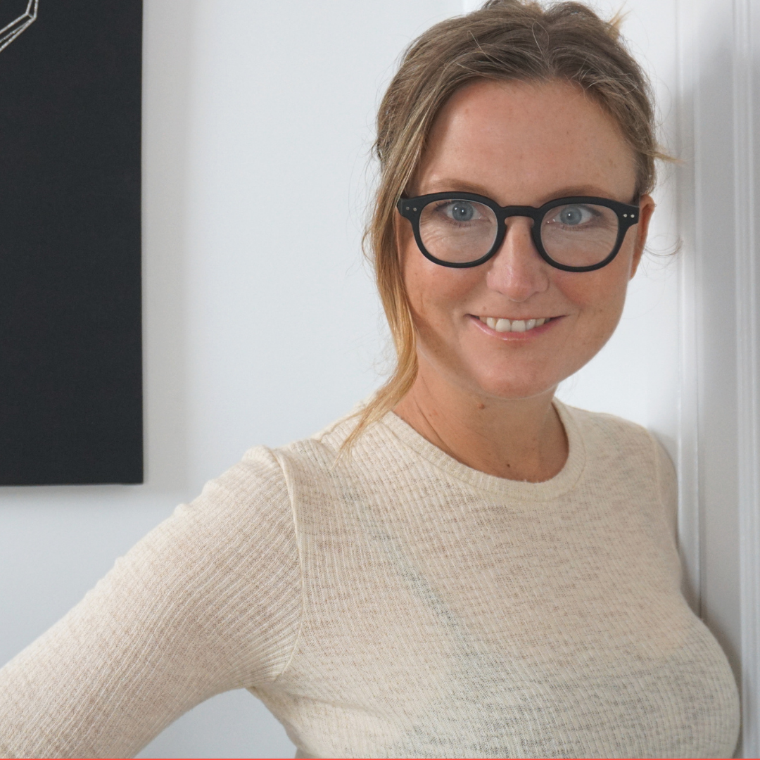 Portrait of Master esthetician and host of Better Skin Workshop. She is leaning up against a white wall looking into the camera. Her skin is clear. Her hair is tied back. She is wearing black framed glasses and a long sleeved white blouse.