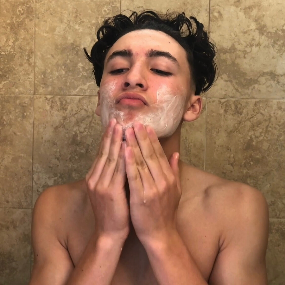 Close up of the face of guy in a bathroom exfoliating  his skin. There is a white product on his face. His hands are on his chin as he is demonstrating how to exfoliate his face.  