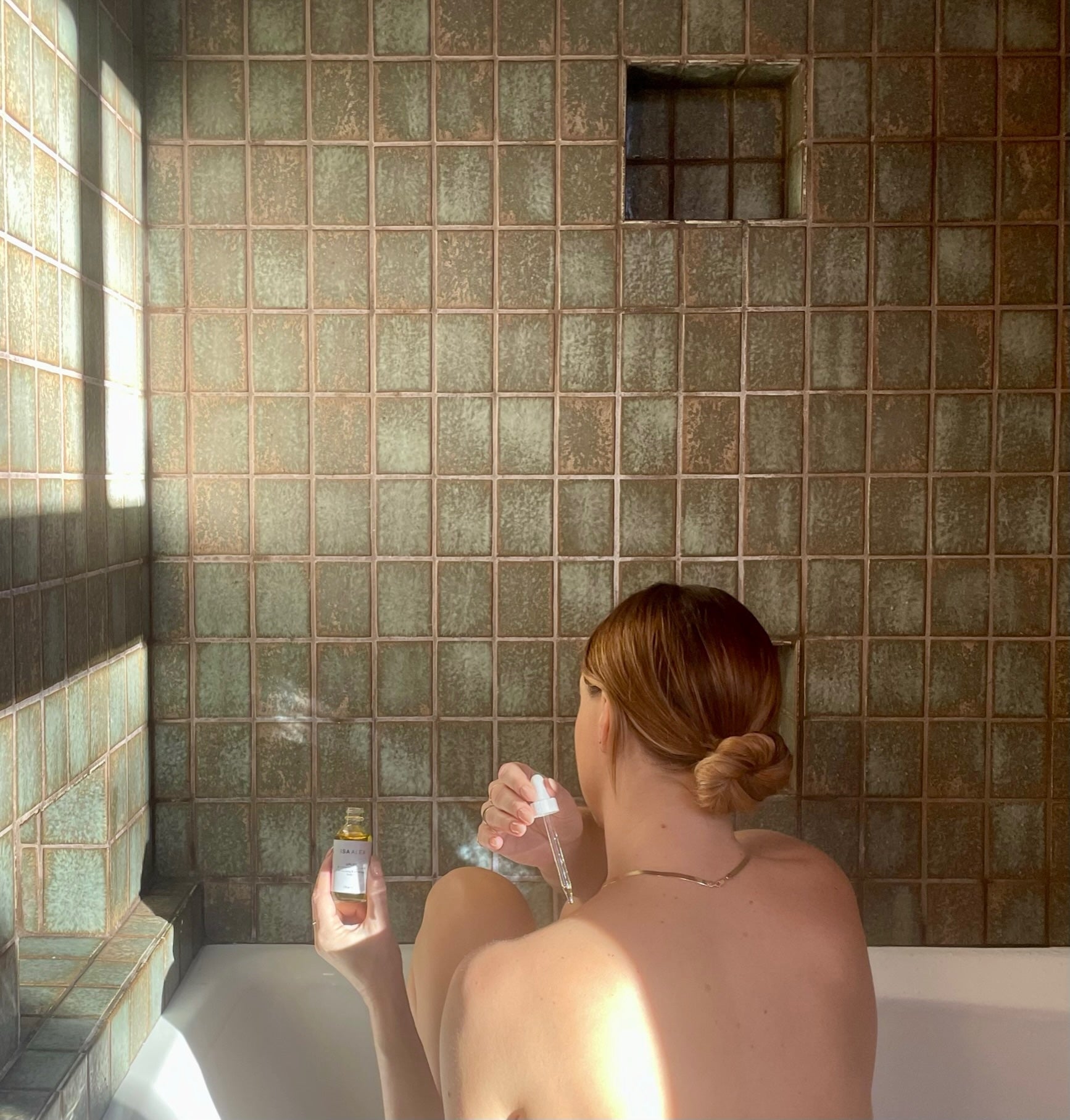 A woman looking into her bathroom mirror as she applies a face oil to her face to do a facial massage. She is wearing a plush white bathrobe. 