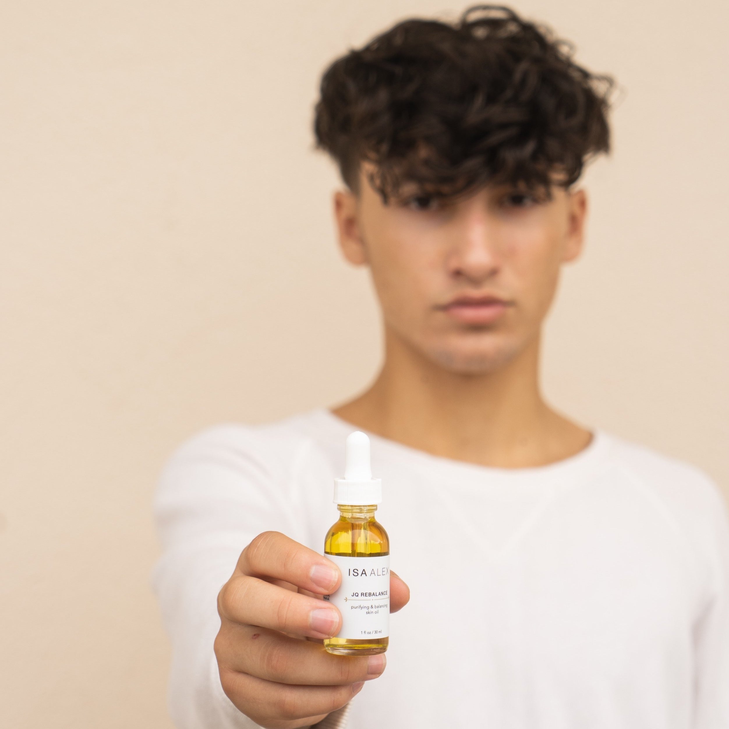 A teen age boy with olive skin, brown hair and dark brown eyes wearing a white long sleeved shirt is seen standing against a light beige colored wall holding out a bottle of skin oil. The bottle is clear with a white label and white dropper so you can see the yellow hue of the skincare oil. The face oil is by ISA ALEX skincare and is called JQ Rebalance Purifying and Rebalancing Face oil. The ambiance of the photo is warm and soft. The teen model is slightly blurred in the background.